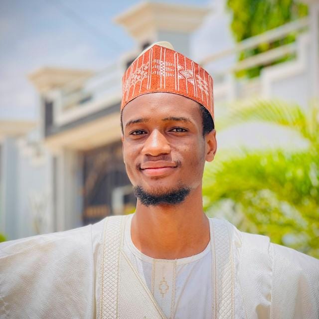 Portrait of Usman Shehu Ahmad smiling, wearing a terracotta embroidered cap and a cream traditional outfit